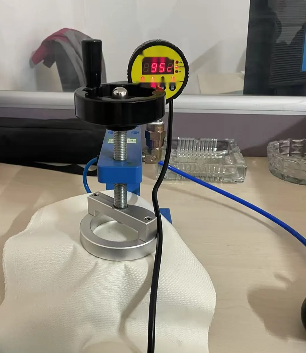 Fabric sample clamped in a screw-down test fixture connected to a digital pressure gauge with a hand pump